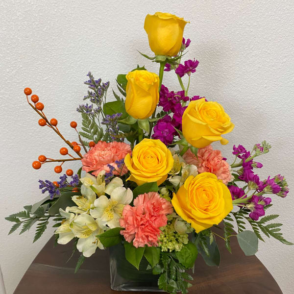 Mixed bouquet of yellow roses, pink carnations, and purple flowers in a glass vase