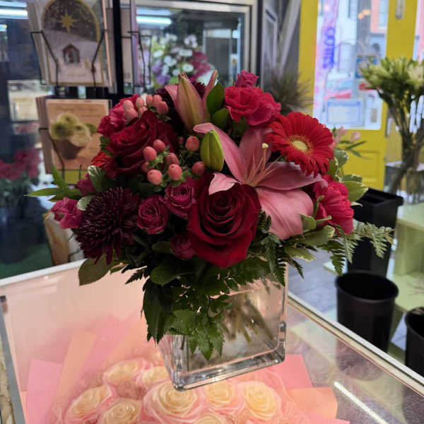 Mixed bouquet of red, pink, and burgundy flowers in a clear square vase