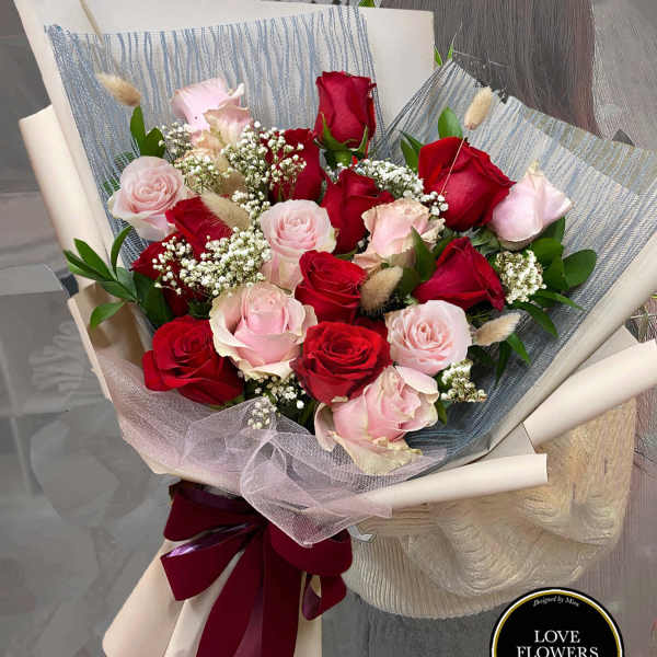 Hand-tied bouquet of red and pink roses with white filler flowers wrapped in cream and gray paper.