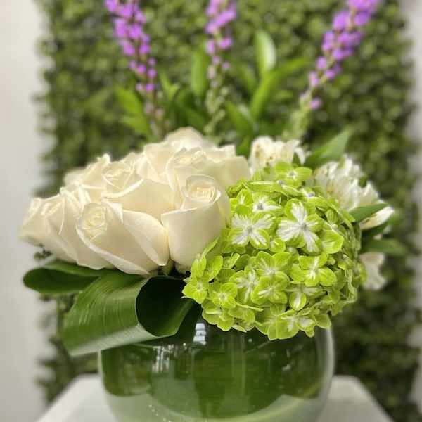 Low arrangement of white roses and green hydrangea in a round green glass vase with tall purple flower spikes.