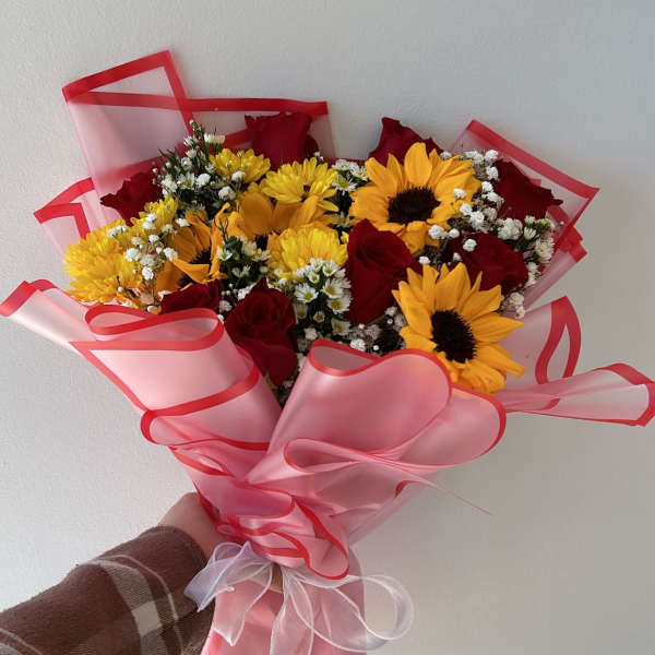 Handheld bouquet of red roses, sunflowers, and small white filler flowers wrapped in pink paper