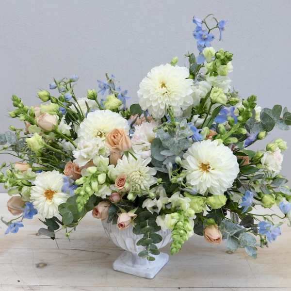 Low floral arrangement in a white pedestal vase with white, peach, and blue blooms