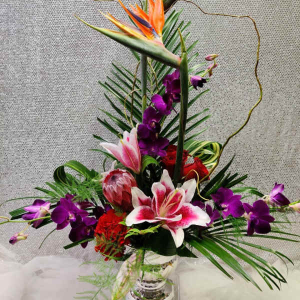 Tall tropical flower arrangement with orange bird of paradise and purple orchids in a glass vase