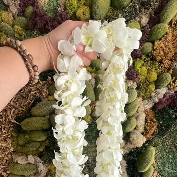 White flower lei held in a hand against a moss wall