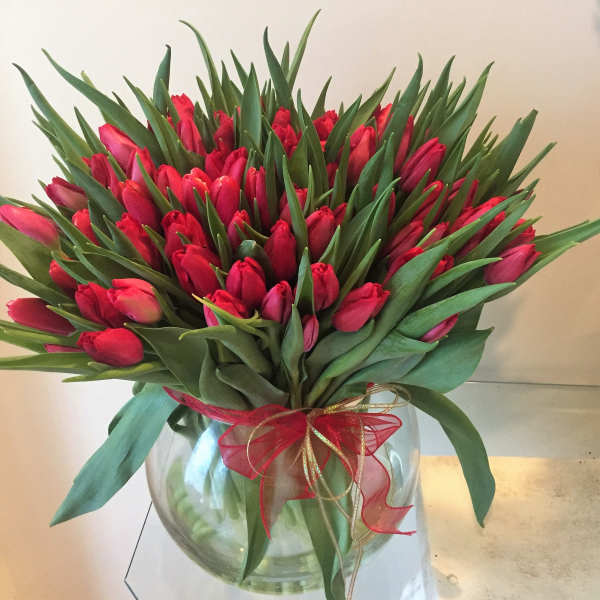 Large bunch of red tulips in a round clear glass vase with a red and gold bow.
