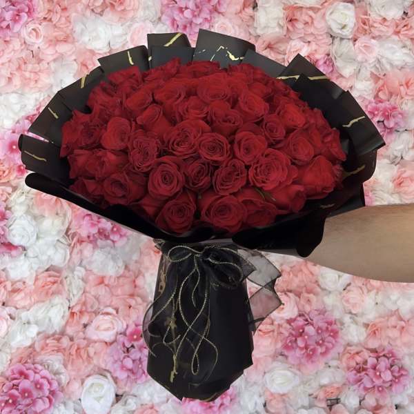 Large bouquet of red roses wrapped in black paper with gold ribbon, held before a pink flower wall