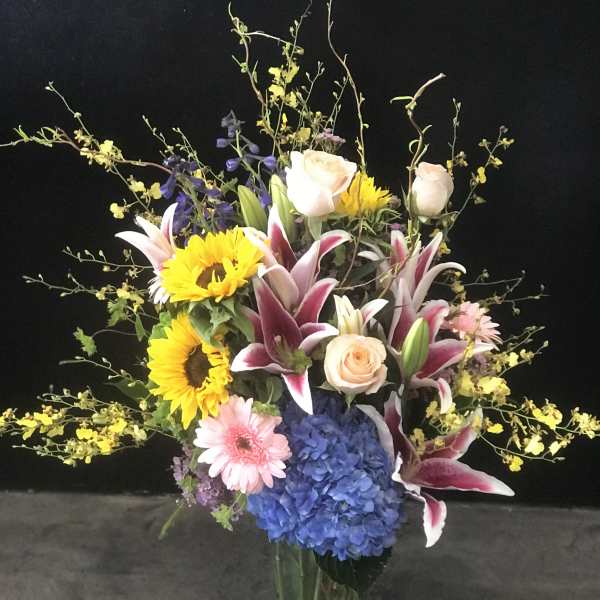 Tall mixed bouquet in a glass vase with sunflowers, lilies, roses, and blue hydrangea