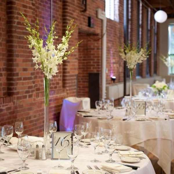 Tall white floral centerpieces on round banquet tables in a brick-walled room