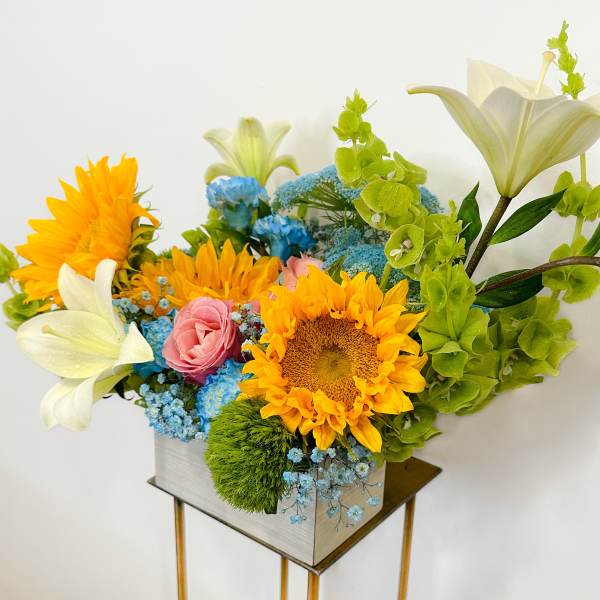 Bright mixed bouquet with sunflowers, lilies, roses, and blue filler flowers in a square vase