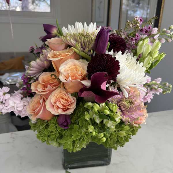 Mixed bouquet of peach roses, white chrysanthemums, and purple flowers in a square vase