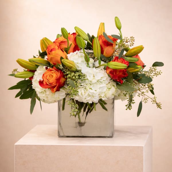 Low glass vase arrangement with orange roses, white hydrangeas, and green lily buds.