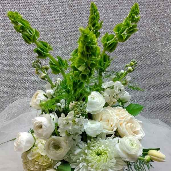Tall white and green flower arrangement with roses, ranunculus, tulips, and Bells of Ireland in a clear glass vase