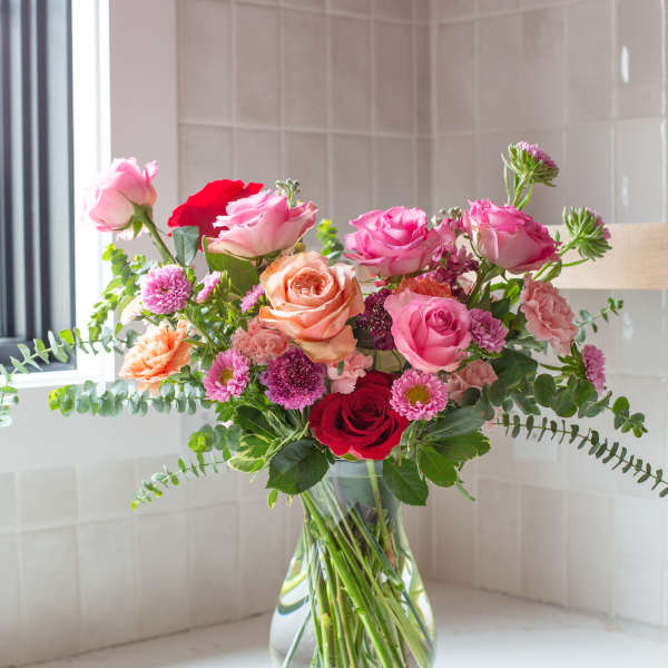 Mixed bouquet of pink, peach, and red roses with small mums in a clear glass vase