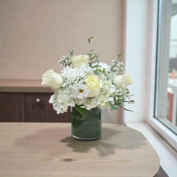 Low arrangement of white and cream roses, hydrangeas, and daisies in a clear glass cylinder vase