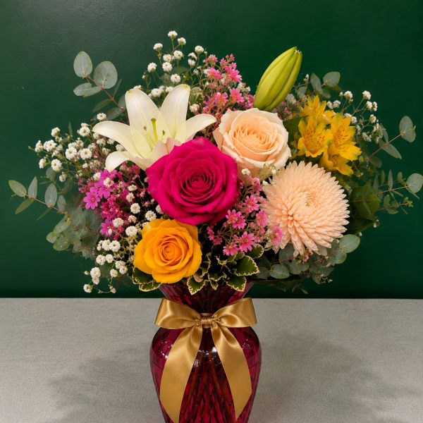 Mixed bouquet of roses, lilies, and daisies in a red vase with a gold ribbon
