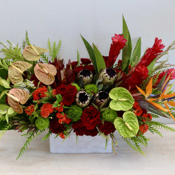 Long centerpiece of red and orange tropical flowers with green and peach anthuriums in a white rectangular vase