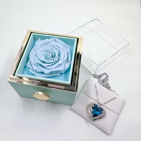 A pale blue rose in a square gift box beside a heart pendant necklace.