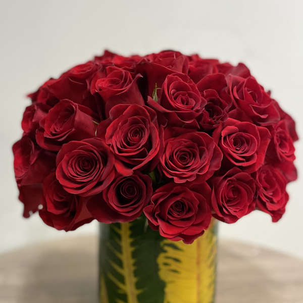 Round arrangement of red roses in a clear glass cylinder vase lined with green and yellow leaves.