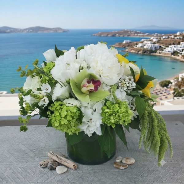 White and green floral arrangement in a dark vase with a seaside view behind it.