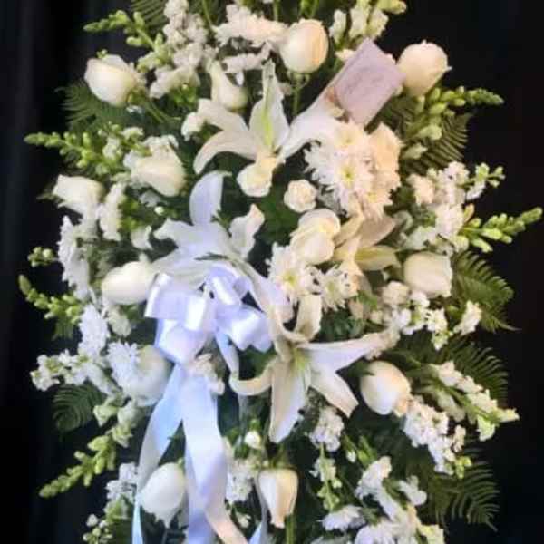 White floral standing spray with lilies, roses, and ribbon on a dark backdrop