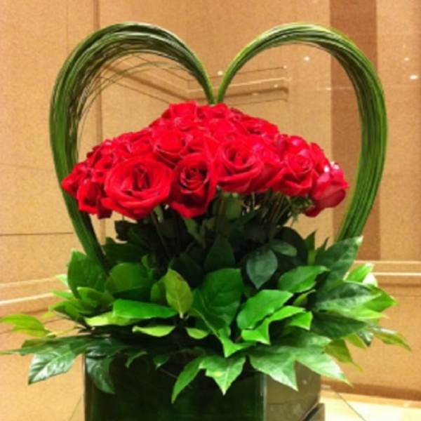 Red roses arranged in a heart-shaped display with green foliage in a square vase