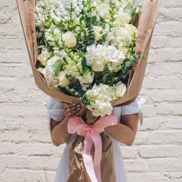 Oversized bouquet of white and cream roses, hydrangeas, and snapdragons wrapped in kraft paper with a pink ribbon.