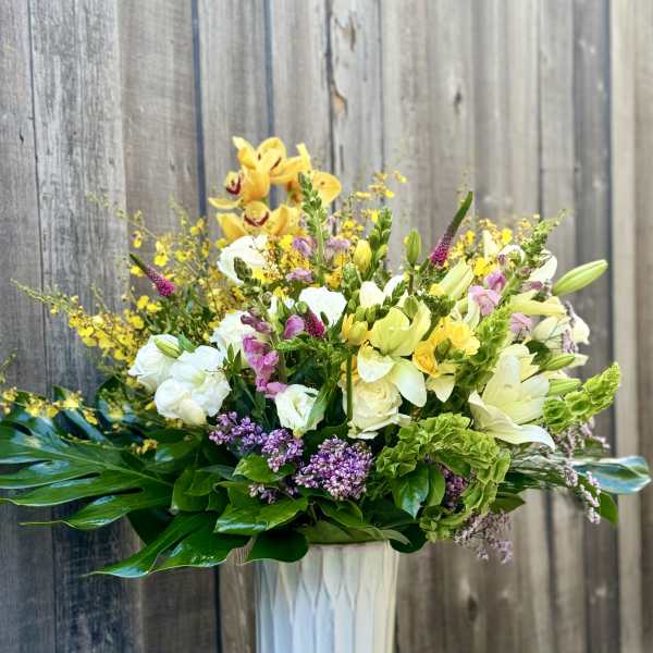 Mixed bouquet in a tall white vase with yellow, white, pink, and purple flowers.