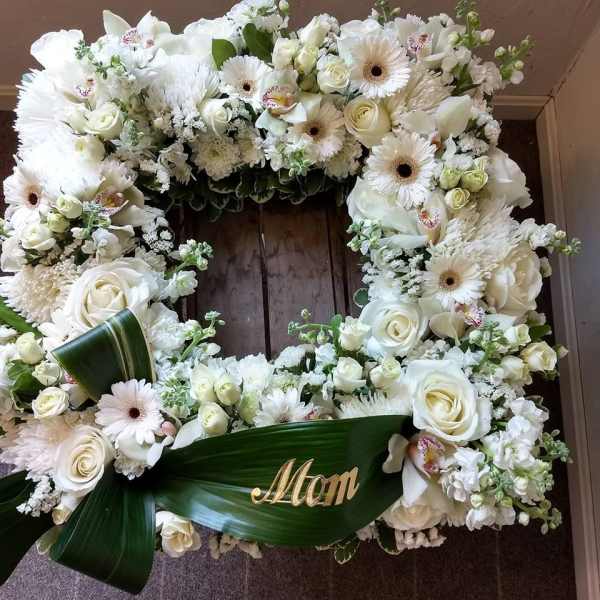 White floral wreath with roses, gerbera daisies, and orchids around a green ribbon marked Mom