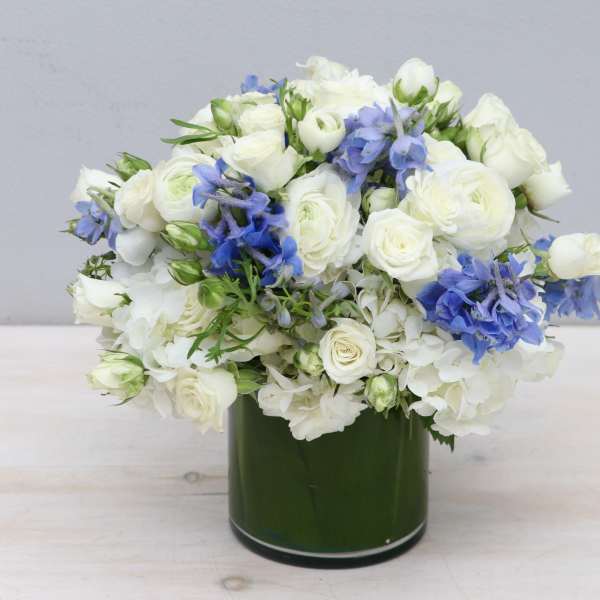 Low round arrangement of white flowers with blue accents in a green cylinder vase