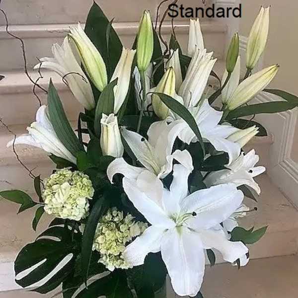 Tall arrangement of white lilies and hydrangeas with monstera leaves in a clear glass vase