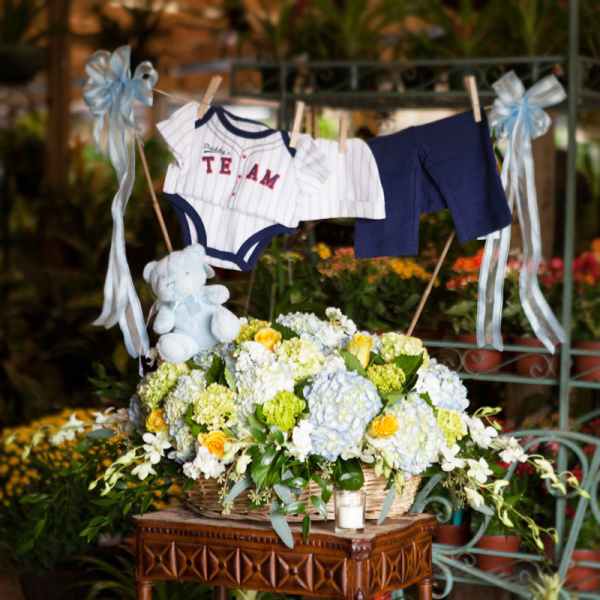 Basket arrangement with blue and white baby clothes and a teddy bear above pastel flowers