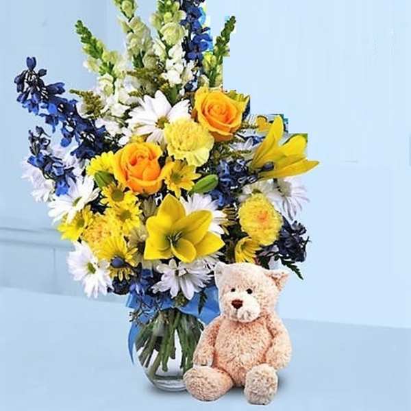 Tall yellow, white, and blue flower arrangement in a vase with a tan teddy bear