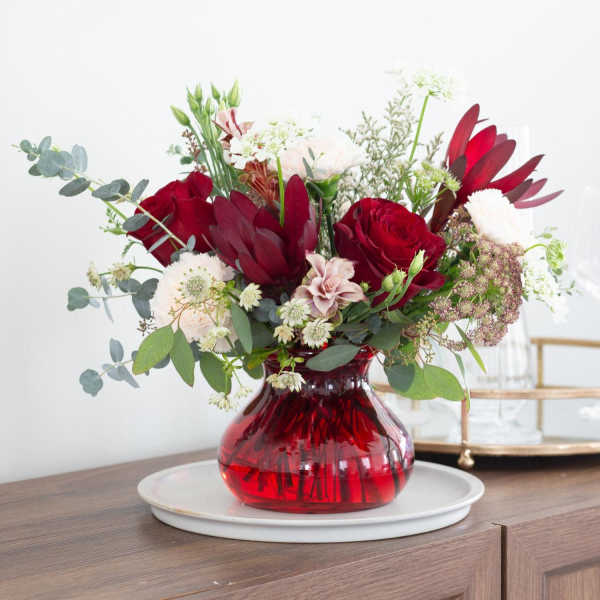 Arrangement of red roses and blush flowers in a round red glass vase on a white plate