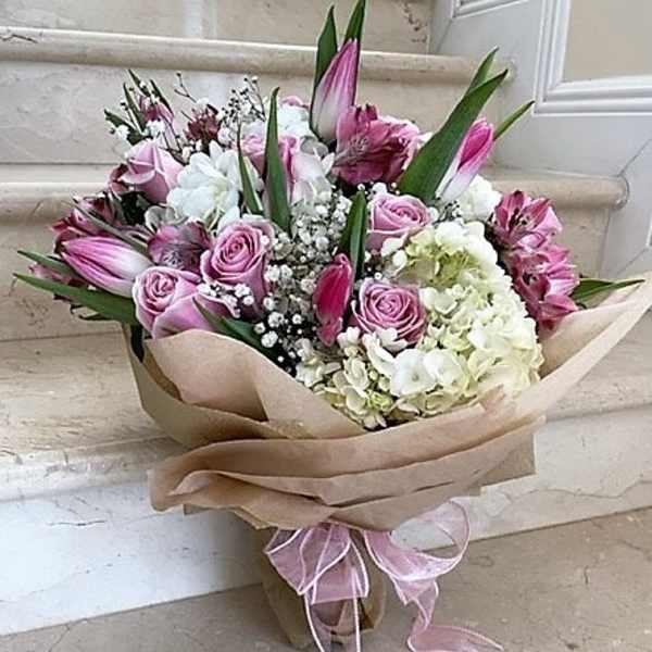 Hand-tied bouquet of pink roses, white hydrangeas, and pink lilies wrapped in kraft paper with a pink ribbon