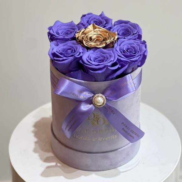 Purple roses in a round lavender box with a gold rose center and ribbon bow