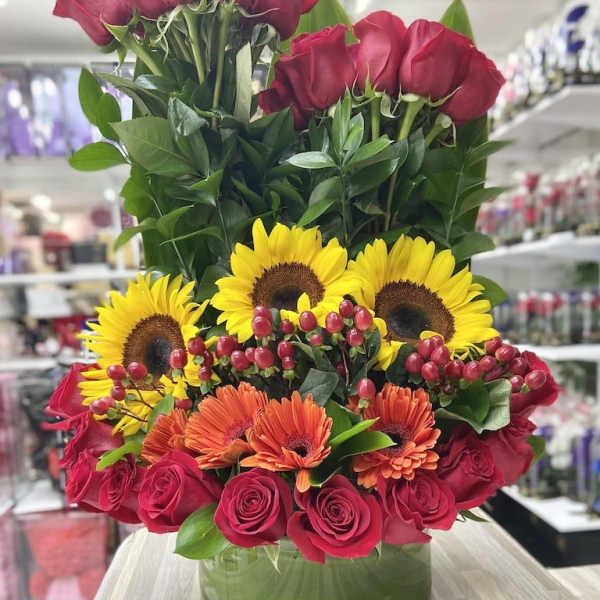 Tall arrangement of red roses, yellow sunflowers, and orange gerbera daisies in a round glass vase.