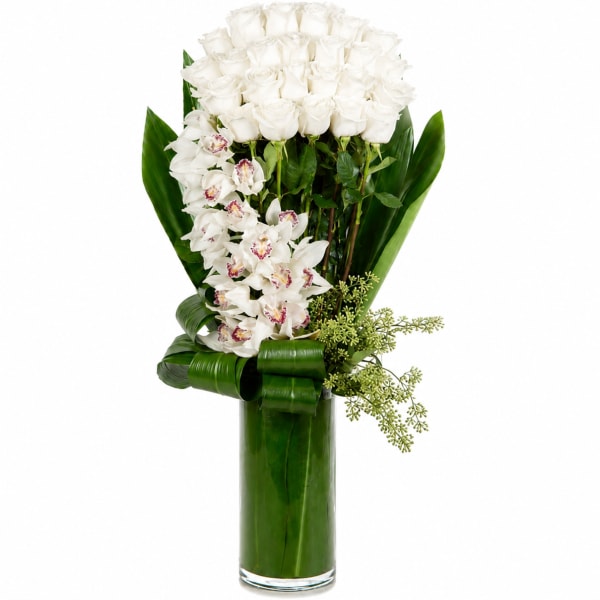 White roses and orchids in a tall glass vase with broad green leaves