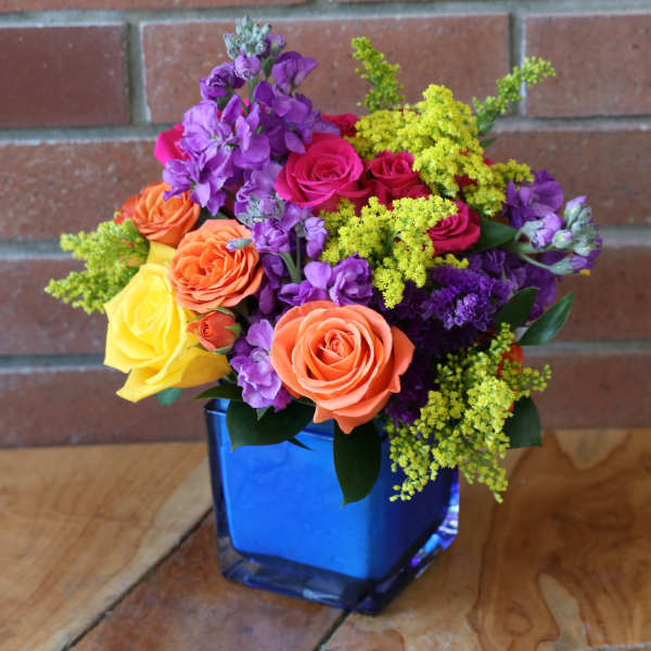 Bright mixed bouquet of orange and yellow roses with purple flowers in a blue glass cube vase