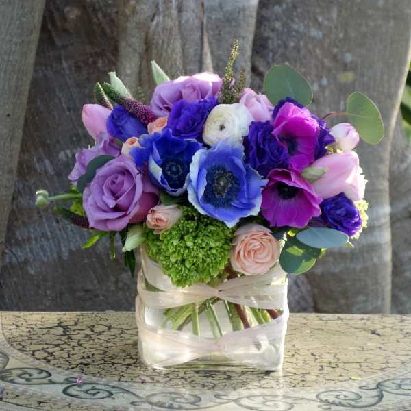 Compact arrangement of purple roses, blue and fuchsia anemones, and peach blooms in a square glass vase.