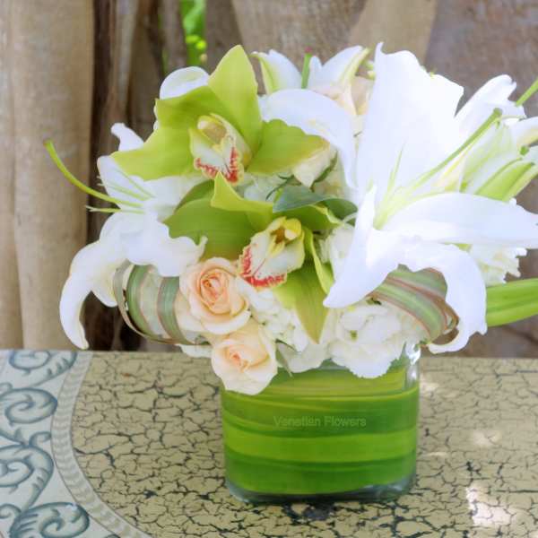 Low arrangement of white lilies, green orchids, and peach roses in a square glass vase