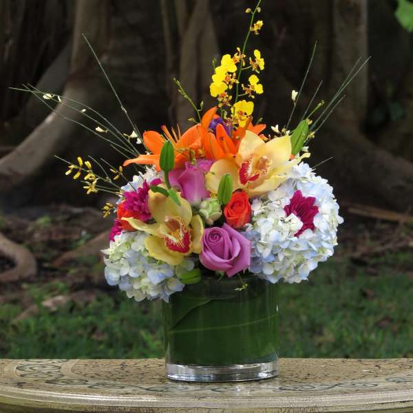 Low glass vase of colorful orchids, lilies, roses, and hydrangeas arranged in a compact mixed bouquet.