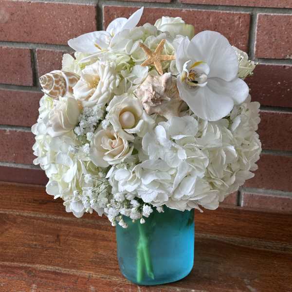 White hydrangeas, roses, and orchids with seashells in a blue glass vase