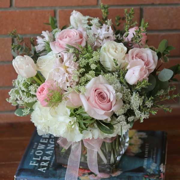 Low pink and white rose arrangement in a clear glass cube vase with ribbon, set on books.