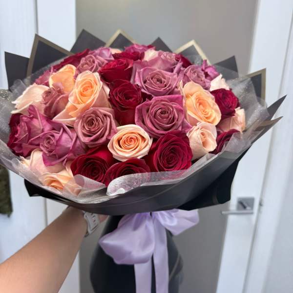 Bouquet of pink, peach, and red roses wrapped in black paper with a lavender ribbon