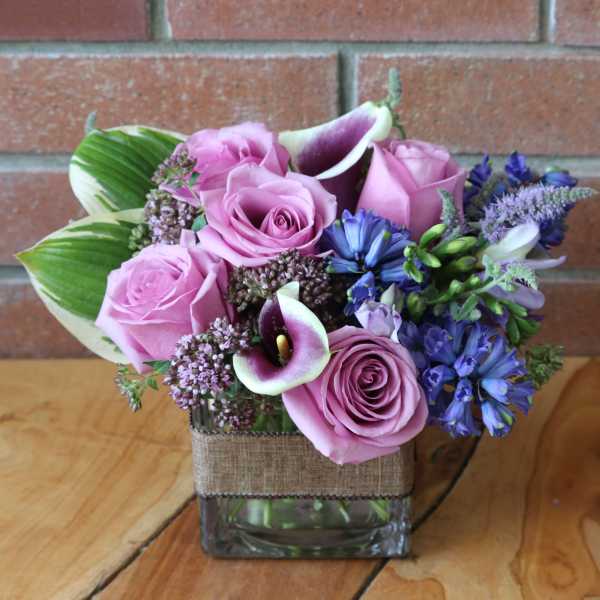 Short arrangement of pink roses, purple calla lilies, and blue flowers in a square glass vase