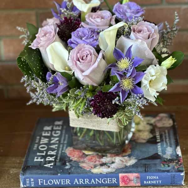 Low lavender and white flower arrangement with roses and calla lilies in a glass cube vase on top of books