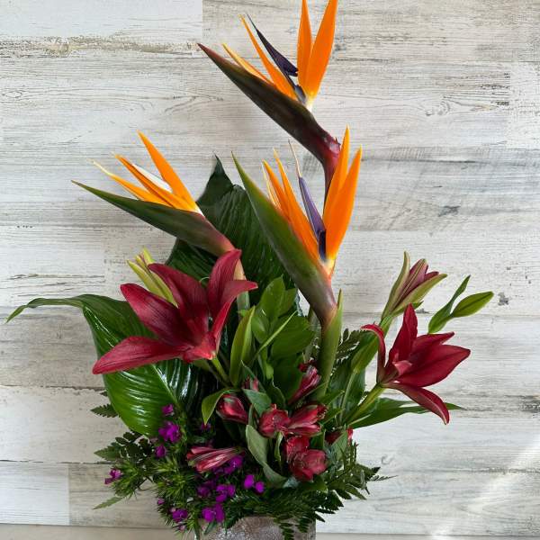 Tropical flower arrangement with orange bird-of-paradise and red lilies in a square vase