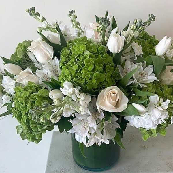 Low arrangement of white roses, tulips, and green hydrangeas in a green glass cylinder vase