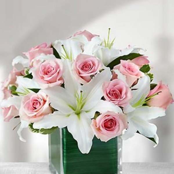 Pink roses and white lilies in a clear glass cube vase lined with a green leaf.