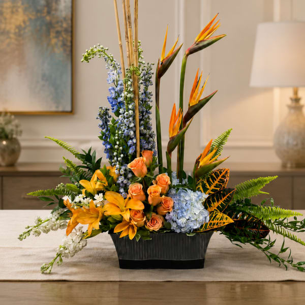 Tropical floral arrangement with orange birds of paradise, roses, lilies, and blue blooms in a black vase